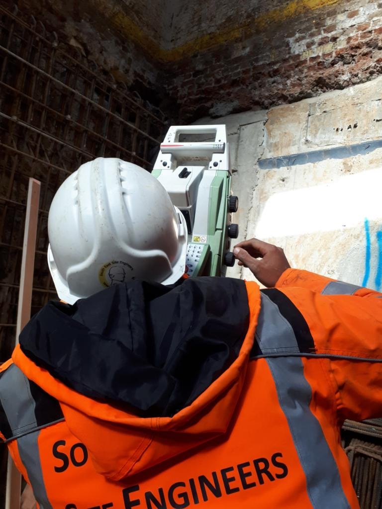 Site engineer in orange jacket operating a Leica total station on an upper floor concrete slab during a London construction project