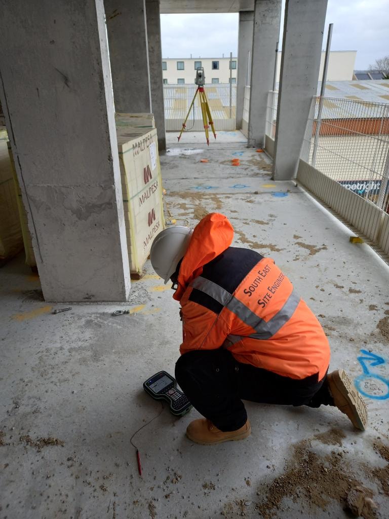 Site engineer in hi-vis jacket using a GPS pole at a London bus depot construction site